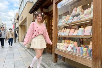 Jeune femme en tenue kawaii pastel avec cardigan rose brodé, jupe plissée crème et chaussures Mary Jane devant une boutique de papeterie japonaise